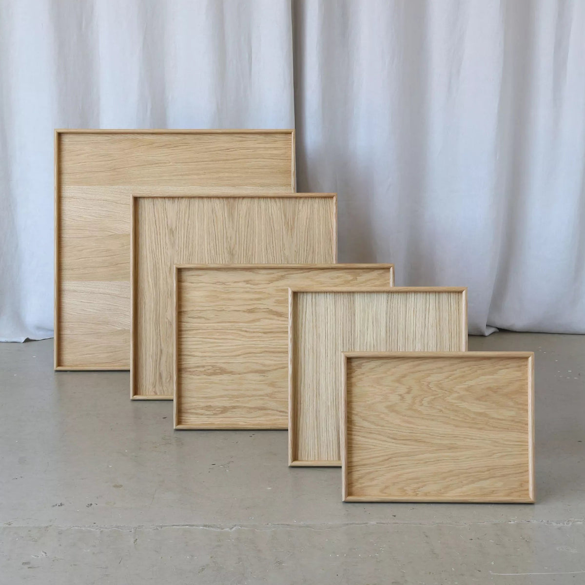 Five rectangular wooden trays in varying sizes and wood grains, arranged in a staggered diagonal row on a concrete floor in front of a white curtain backdrop.
