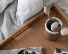 Top-down angle of a wooden tray on a bed, displaying a ceramic vase with dried flowers and a cup of coffee.