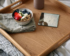 Overhead view of a wooden ottoman tray, holding a breakfast bowl, a textured napkin, and a decorative mirror coaster.