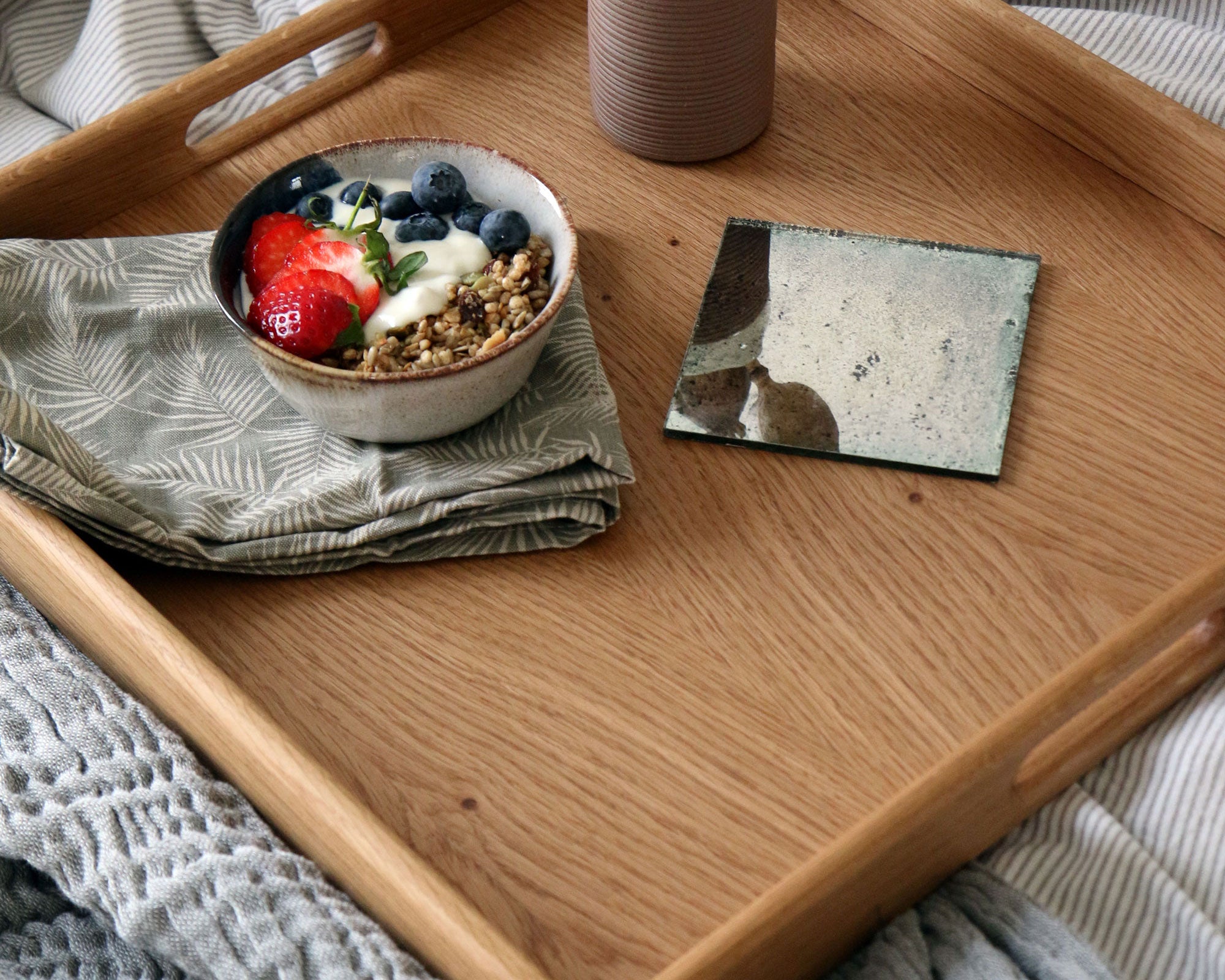 Overhead view of a wooden ottoman tray, holding a breakfast bowl, a textured napkin, and a decorative mirror coaster.