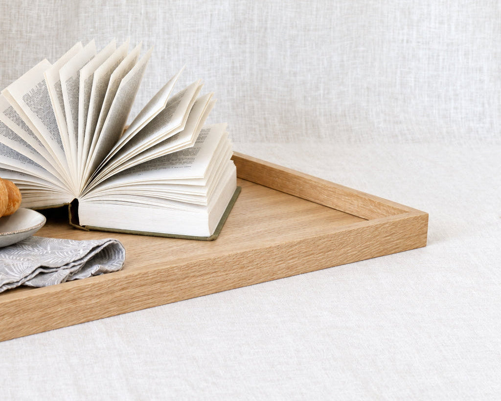 A light natural wood tray holding an open book and a folded gray cloth, set against a soft white linen backdrop.