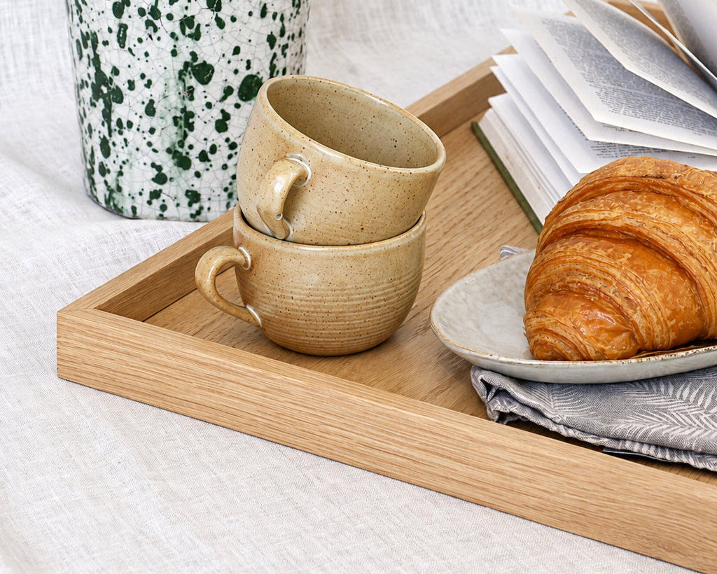 A styled breakfast scene on a light wood tray with two stacked beige ceramic mugs, an open book, a croissant on a small plate, and a tall white speckled planter with green leaves against a white linen backdrop.