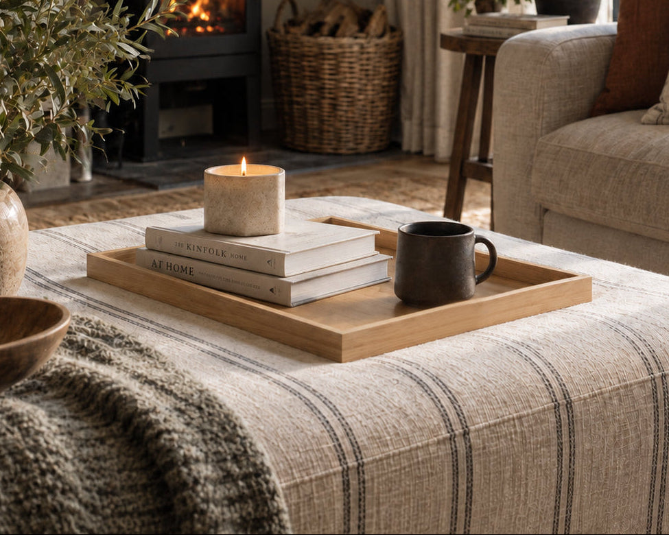 A cozy living room scene with a black wood-burning stove, beige sofa, striped throw blanket, and a wooden tray holding stacked books and a candle on the ottoman.