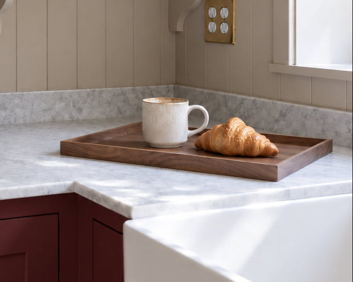 A styled kitchen corner with cream paneled walls, open floating shelves holding bowls, plates, and plants, a white marble countertop, dark red cabinets, and a window with natural light. A walnut tray on the counter holds a white mug and a croissant.