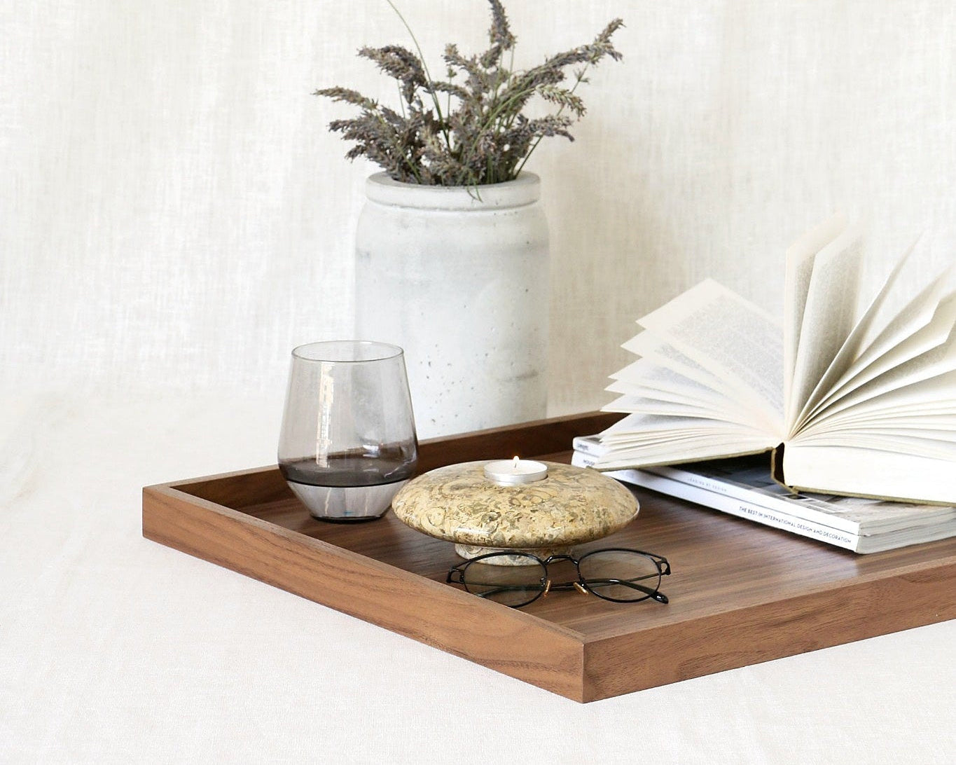 A rectangular wooden serving tray styled with an open book, a small glass, a round stone candle holder, and a pair of eyeglasses, set against a neutral white linen backdrop with a ceramic vase of dried lavender stems.