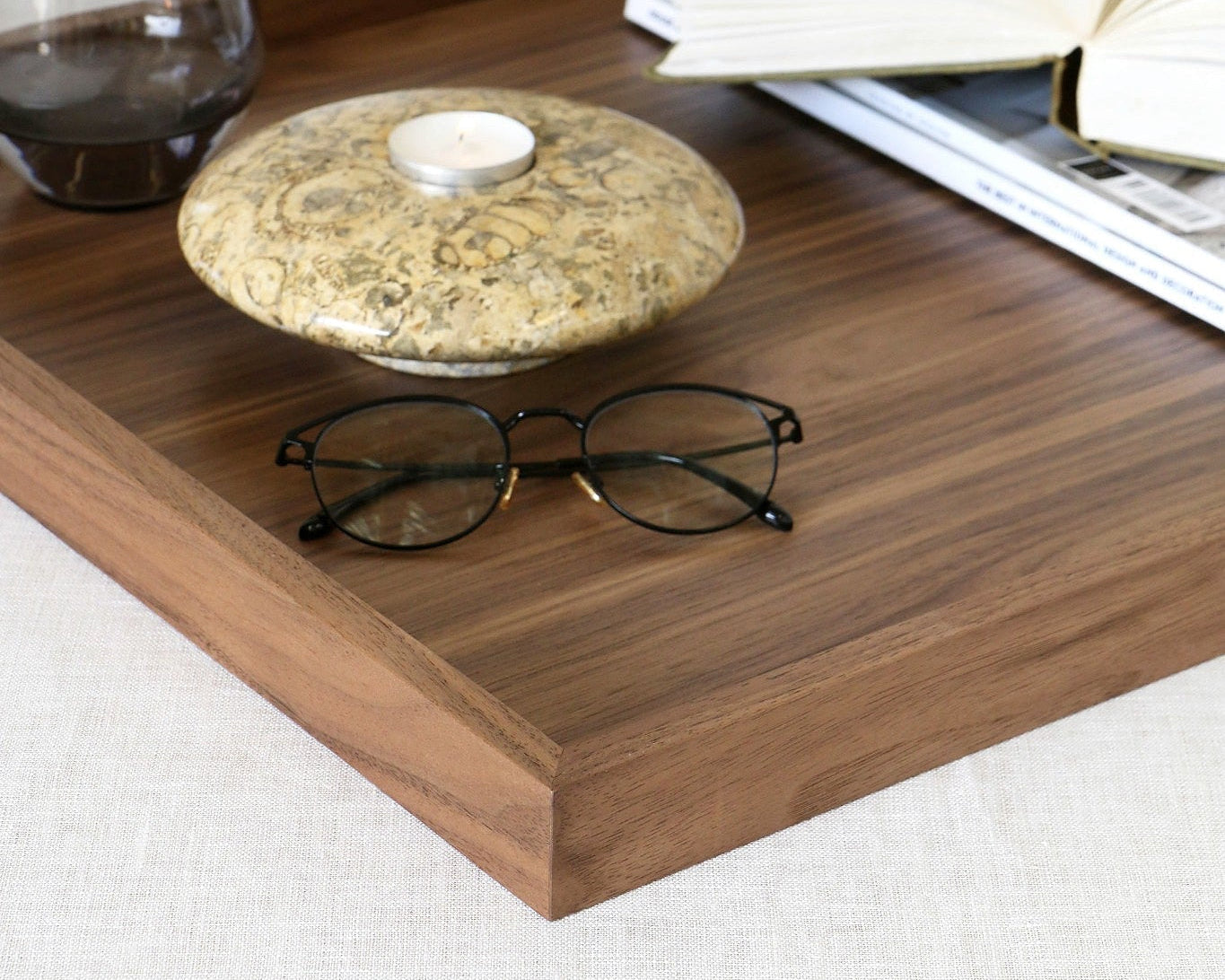 A rectangular wooden tray with straight corners sits on a light fabric surface, styled with an open book, a folded magazine, a small round stone holder with a candle, and a dark glass cup.