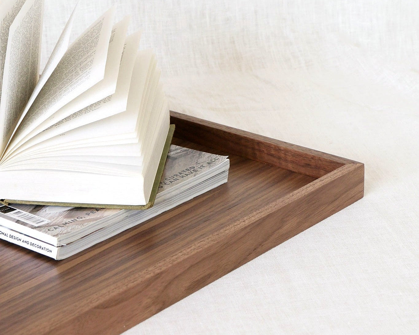 A rectangular wooden tray with a warm medium-brown finish, styled with an open book and a magazine on a light beige textured fabric surface.