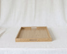 A rectangular light oak wood serving tray with two cutout handles, displayed on a draped white fabric background.