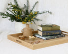 A light natural oak serving tray with cutout handles, styled with a small ceramic vase of yellow flowers, a small brown jug, and a stack of books on a white tablecloth against a white linen background.