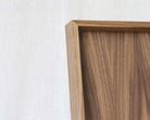 Close-up of a wooden tray frame corner with a warm medium-brown wood grain against a light beige textured wall.