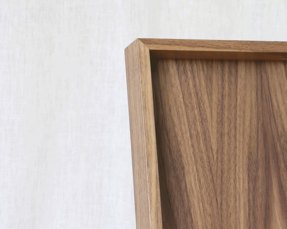 Close-up of a wooden tray frame corner with a warm medium-brown wood grain against a light beige textured wall.