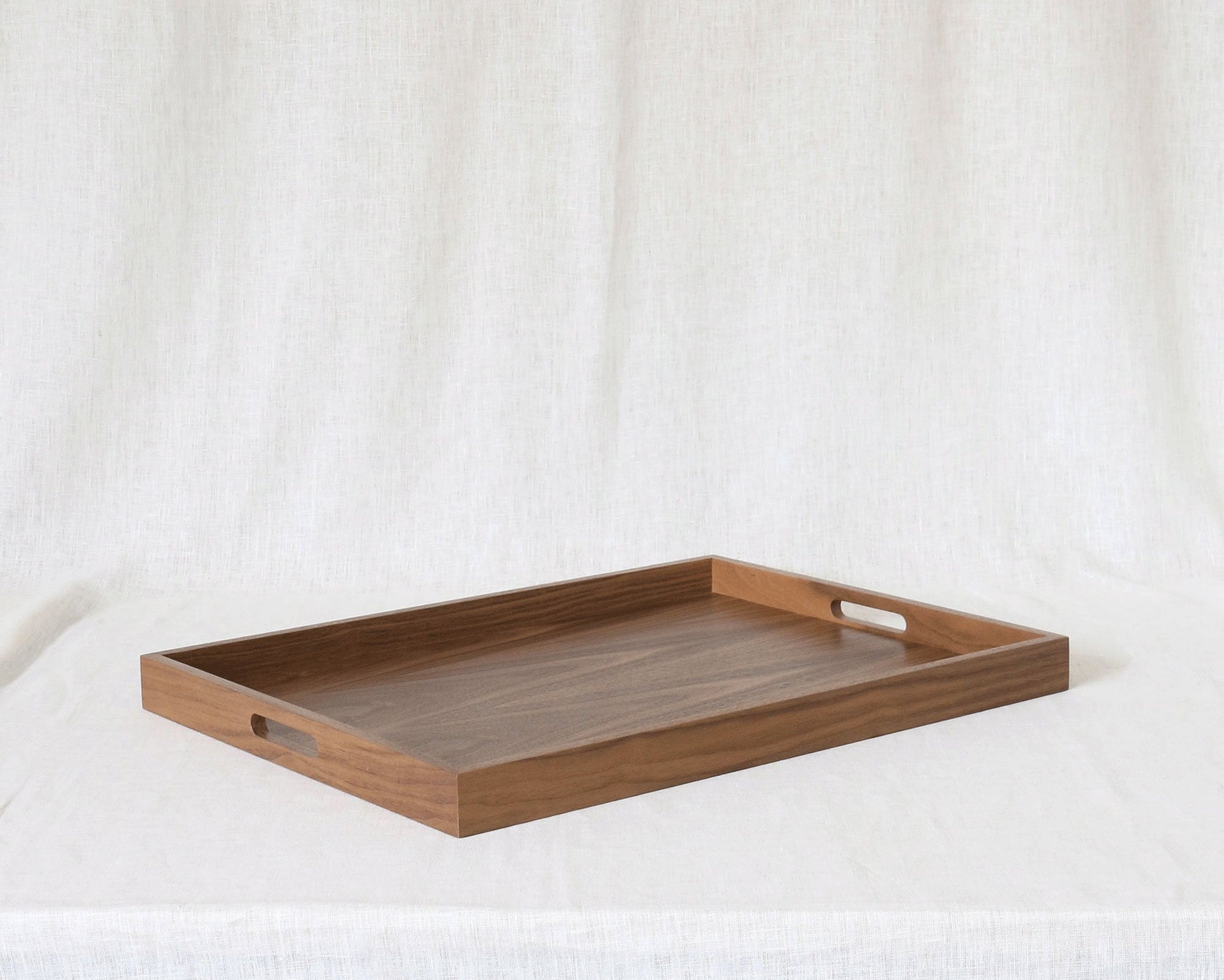 A rectangular wooden serving tray with two cutout handles, resting on a draped white fabric background.