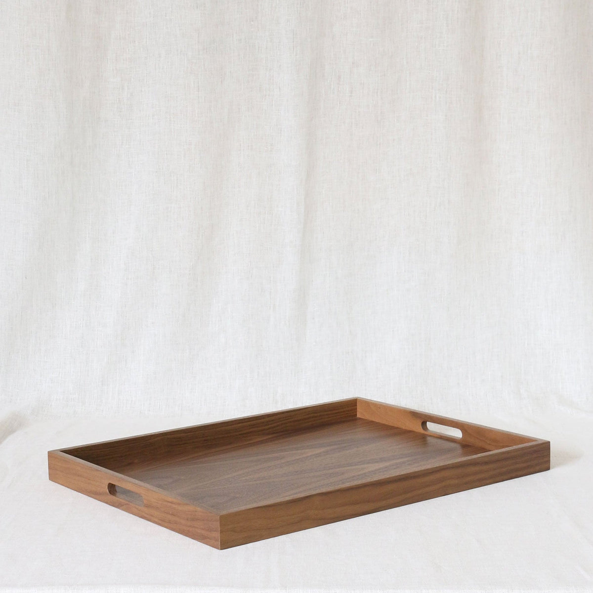 A rectangular wooden serving tray with two cutout handles on the sides, shown on a draped white fabric background.