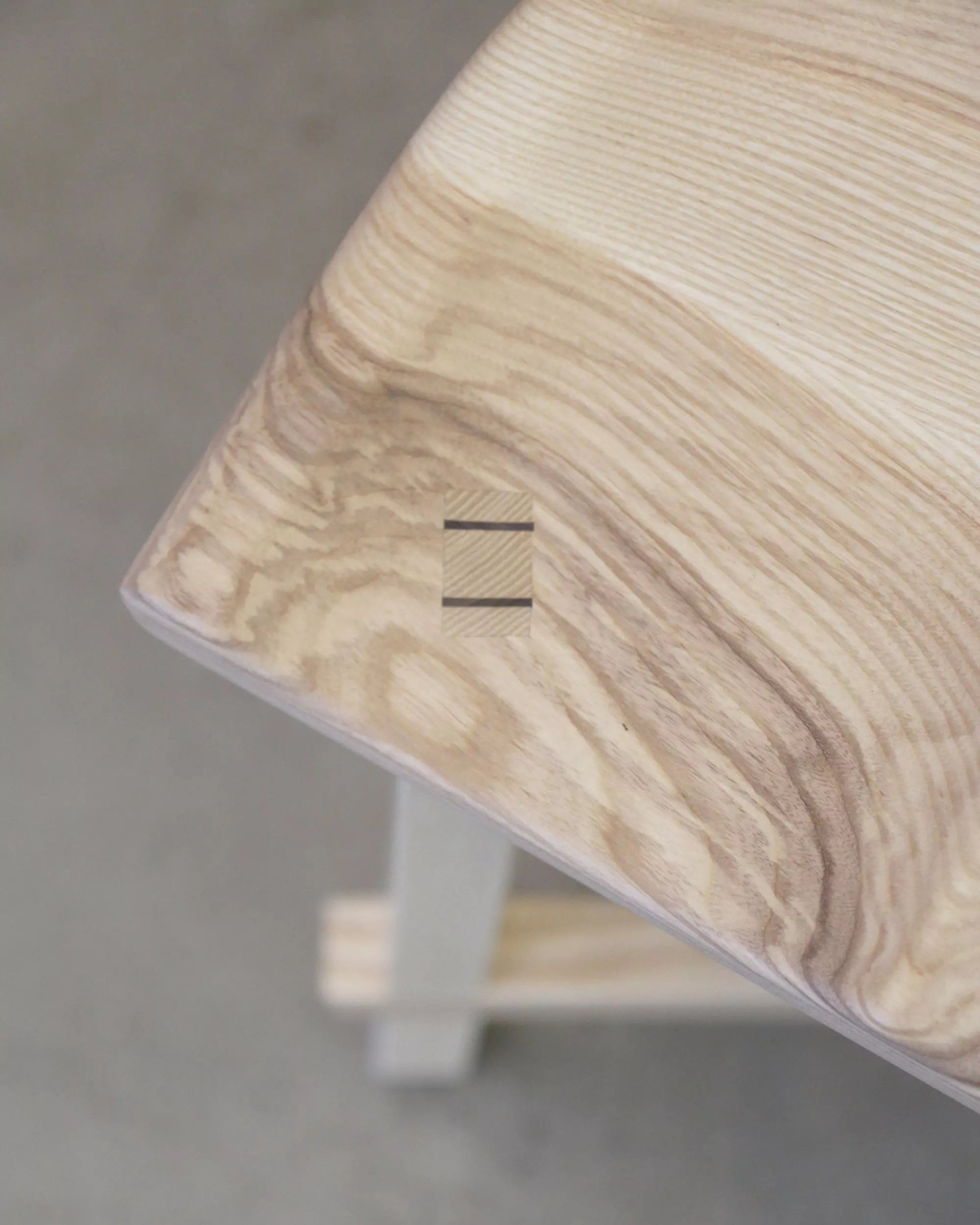 Close-up of a wooden stool with a carved seat joinery detail on a grey floor.