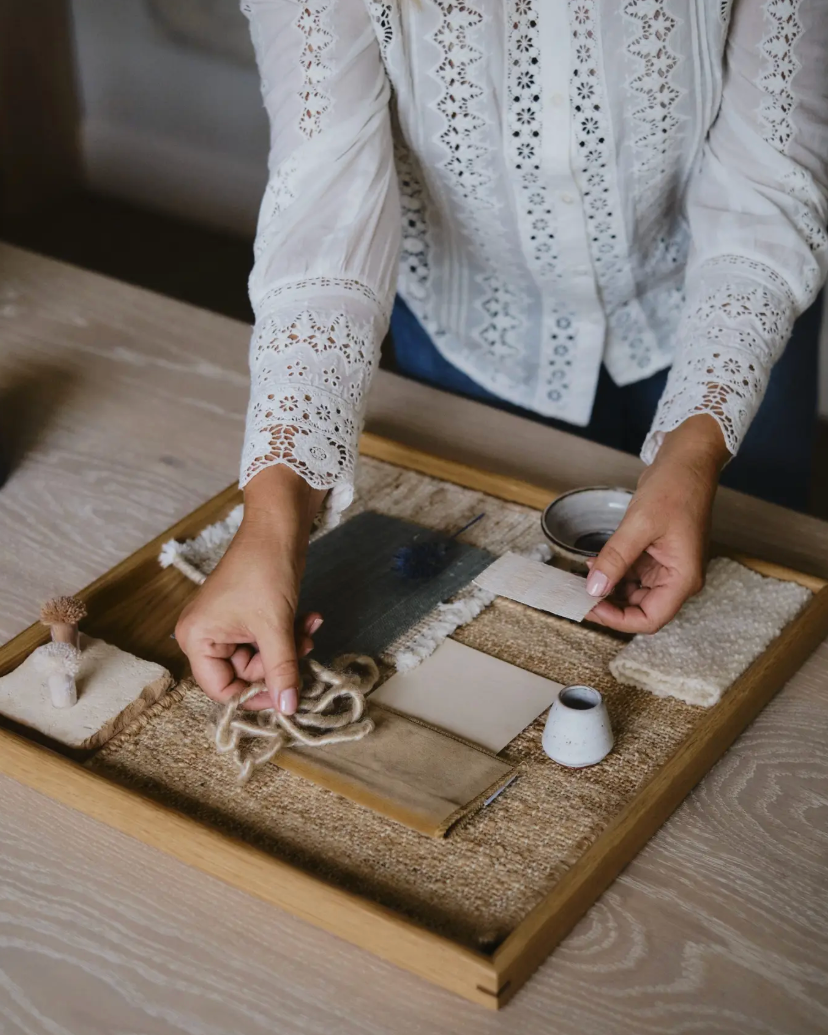 A person wearing a white lace-trim blouse is arranging craft or decor items on a wooden tray over a textured beige surface. Visible items include small fabric pieces, rope, a small white cup, and a dark rectangular object in a styled indoor setting.