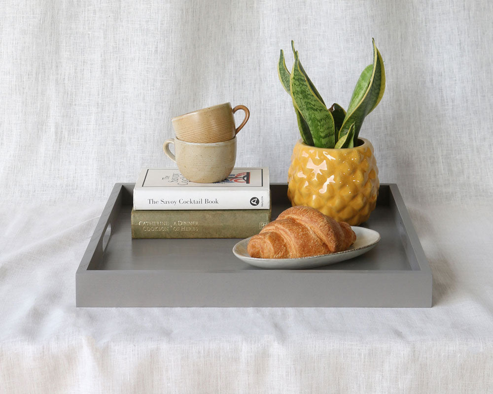Grey ottoman tray featuring a yellow pineapple-style planter, croissant, stacked mugs, and books.