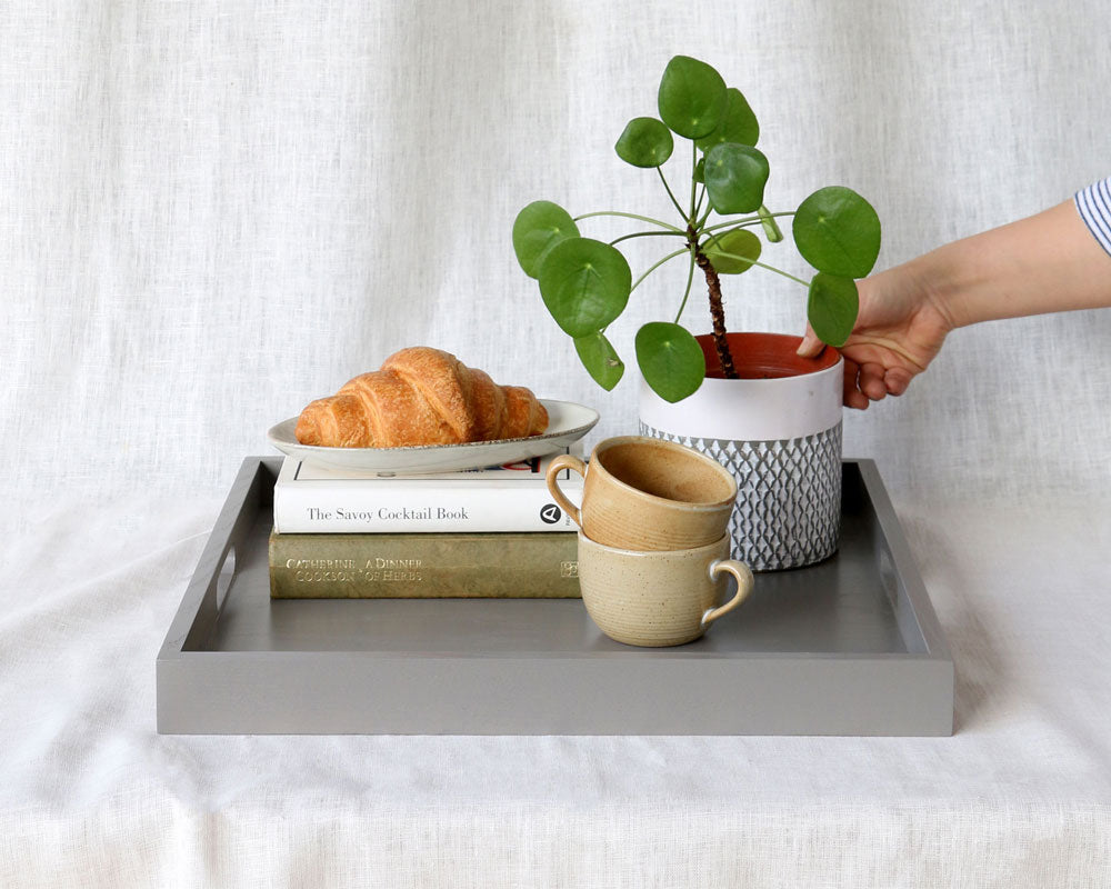 Grey ottoman tray styled with a plant, croissant, ceramic mugs, and books, with a hand placing the plant.