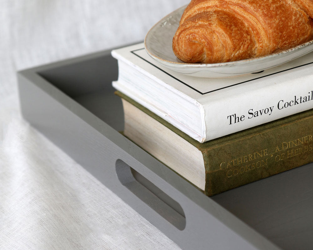 Grey ottoman tray styled with a croissant on a plate and stacked books including “The Savoy Cocktail Book.”