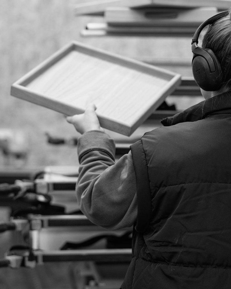 A craftsman wearing headphones and a padded vest inspects a handcrafted wooden tray in a woodworking studio, with neatly stacked trays in the background.