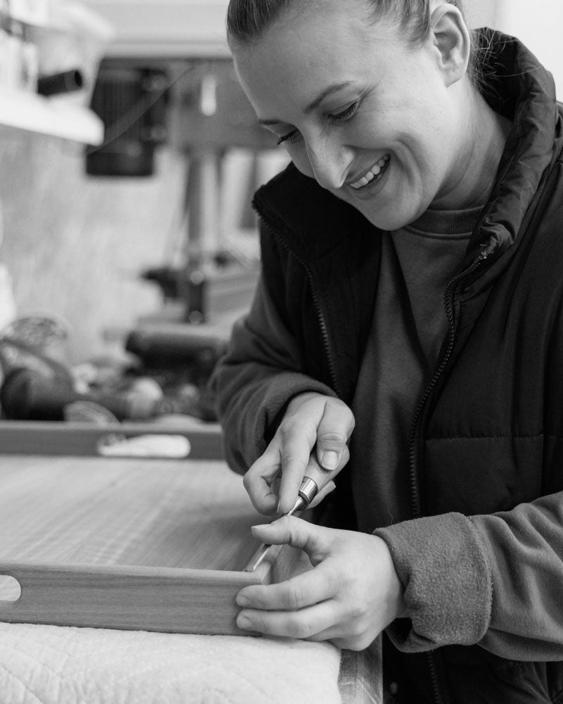 A smiling craftswoman carefully carves details into the edge of a wooden tray using a chisel, showcasing precise handcrafting skills.