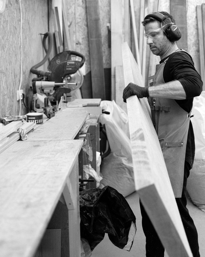 A woodworker wearing safety goggles, earmuffs, and gloves lifts a large wooden plank in a workshop, preparing it for cutting.