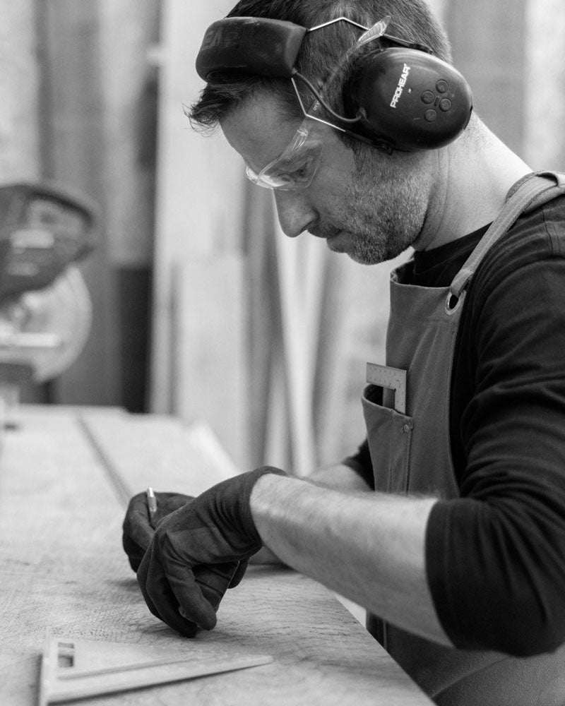 A focused woodworker wearing safety glasses, earmuffs, and gloves meticulously marks a piece of wood in a workshop, preparing for precise cuts.