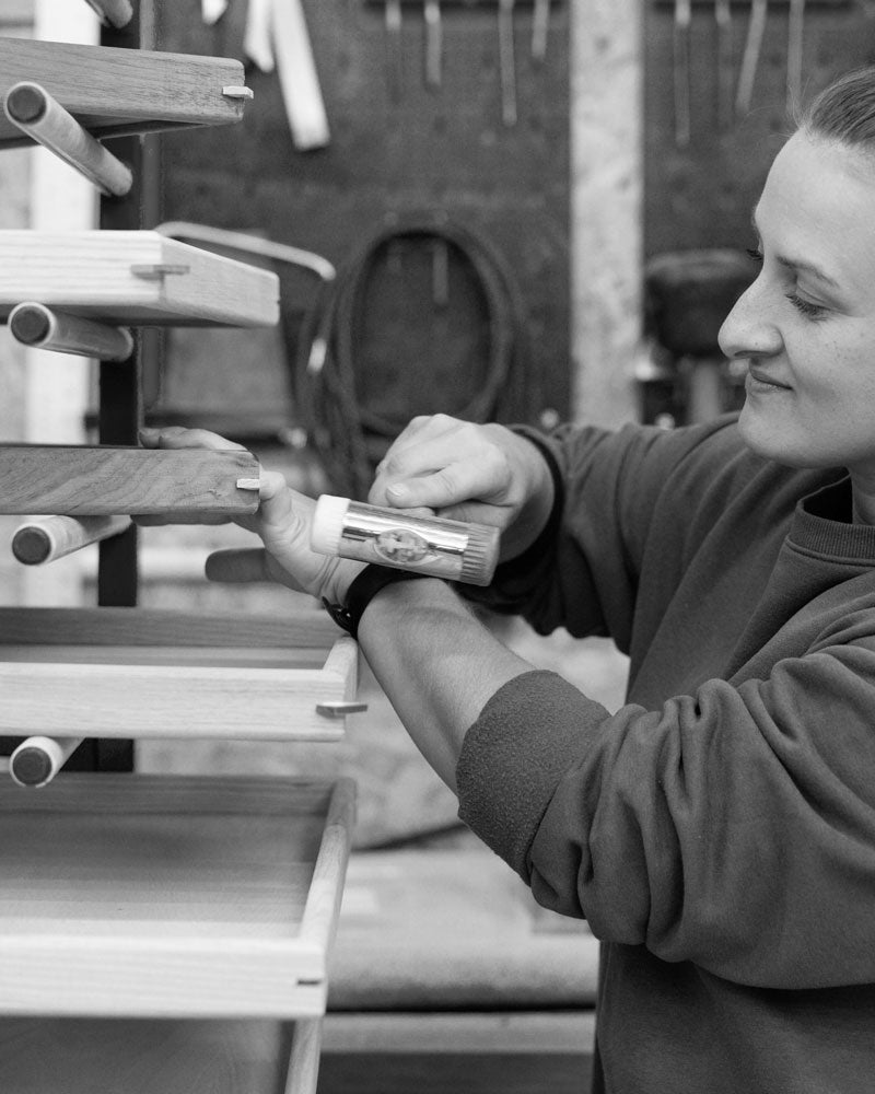 A craftswoman in a workshop gently taps a wooden tray with a hammer, adjusting it on a drying rack with trays stacked on wooden dowels in the background