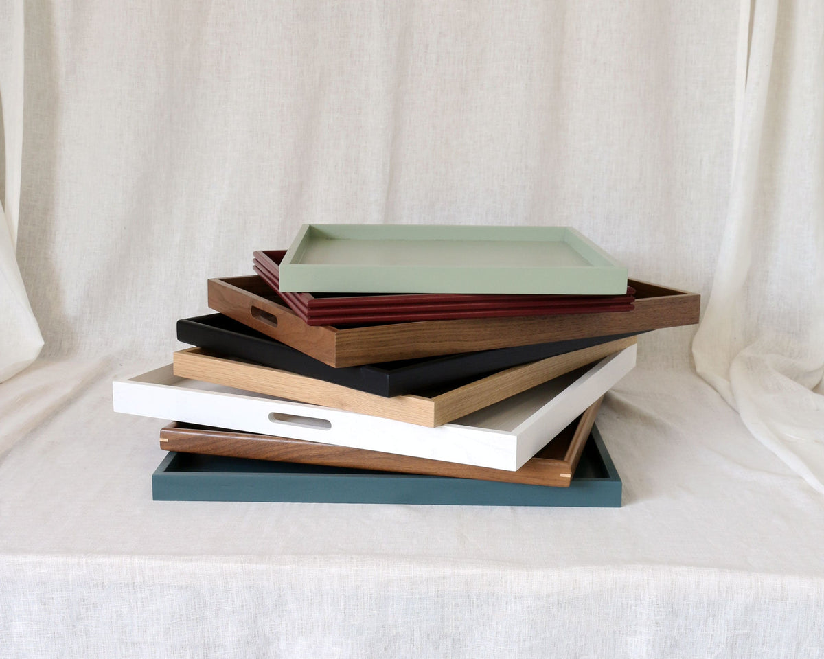 A stack of rectangular serving trays in various colors, including sage green, brown, black, white, beige, and blue, displayed on a draped white fabric backdrop.