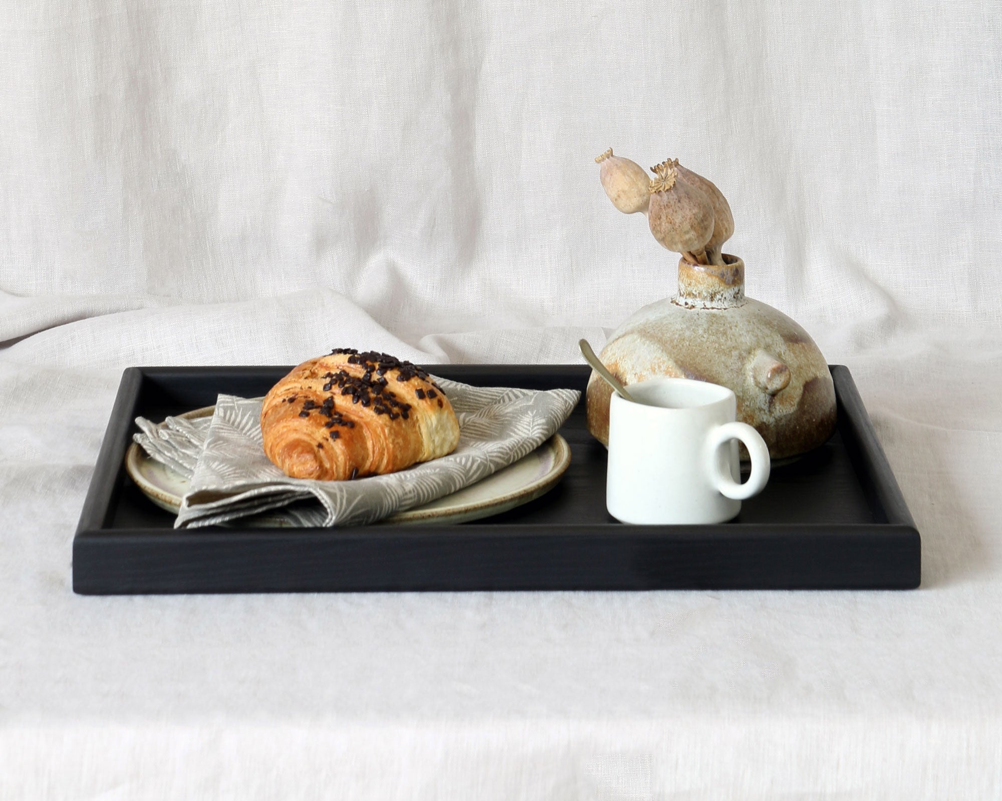 Wooden tray for footstool featuring a black handcrafted design, elegantly styled with a breakfast setting including a croissant, a white ceramic cup, and a rustic decorative vase.