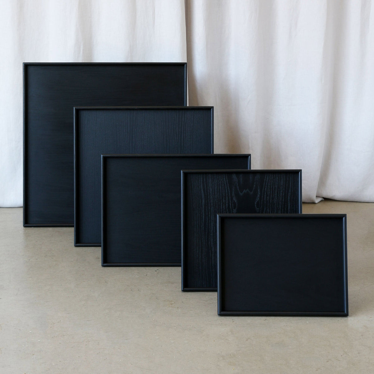 Five black rectangular trays of varying sizes and grain patterns are arranged in a staggered row on a light beige floor in front of a draped off-white fabric backdrop.