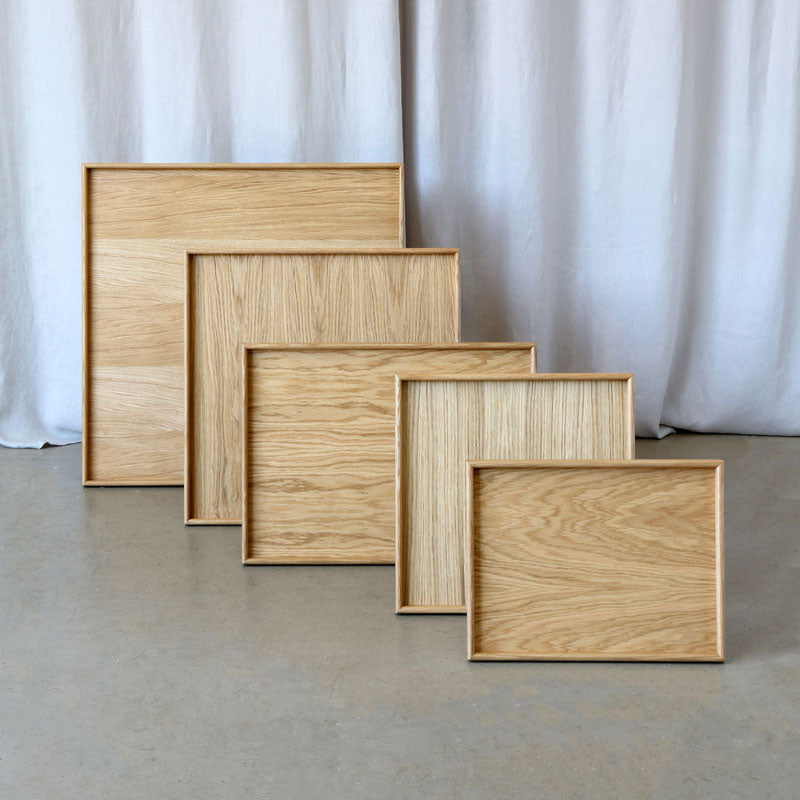 A set of five wooden trays in different sizes, made from light oak wood with a natural finish. The trays have a minimalist design with raised edges and are displayed in a staggered arrangement against a neutral backdrop with a light-colored curtain.