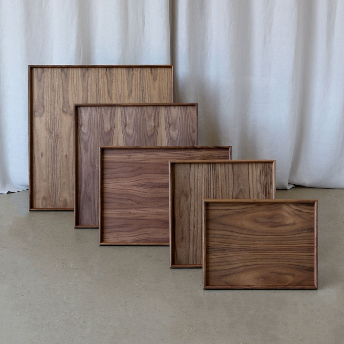 Five rectangular wooden picture frames with varying sizes and grain patterns, arranged in a staggered row on a concrete floor in front of a draped off-white fabric backdrop.