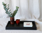 A decorative tray in black, adorned with eucalyptus stems in a ceramic vase, a red cup, and a book, displayed on a soft fabric surface.