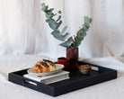 A coffee table tray in black holding a plate with a chocolate croissant, a stack of books, and decorative ceramic items, styled with eucalyptus stems.