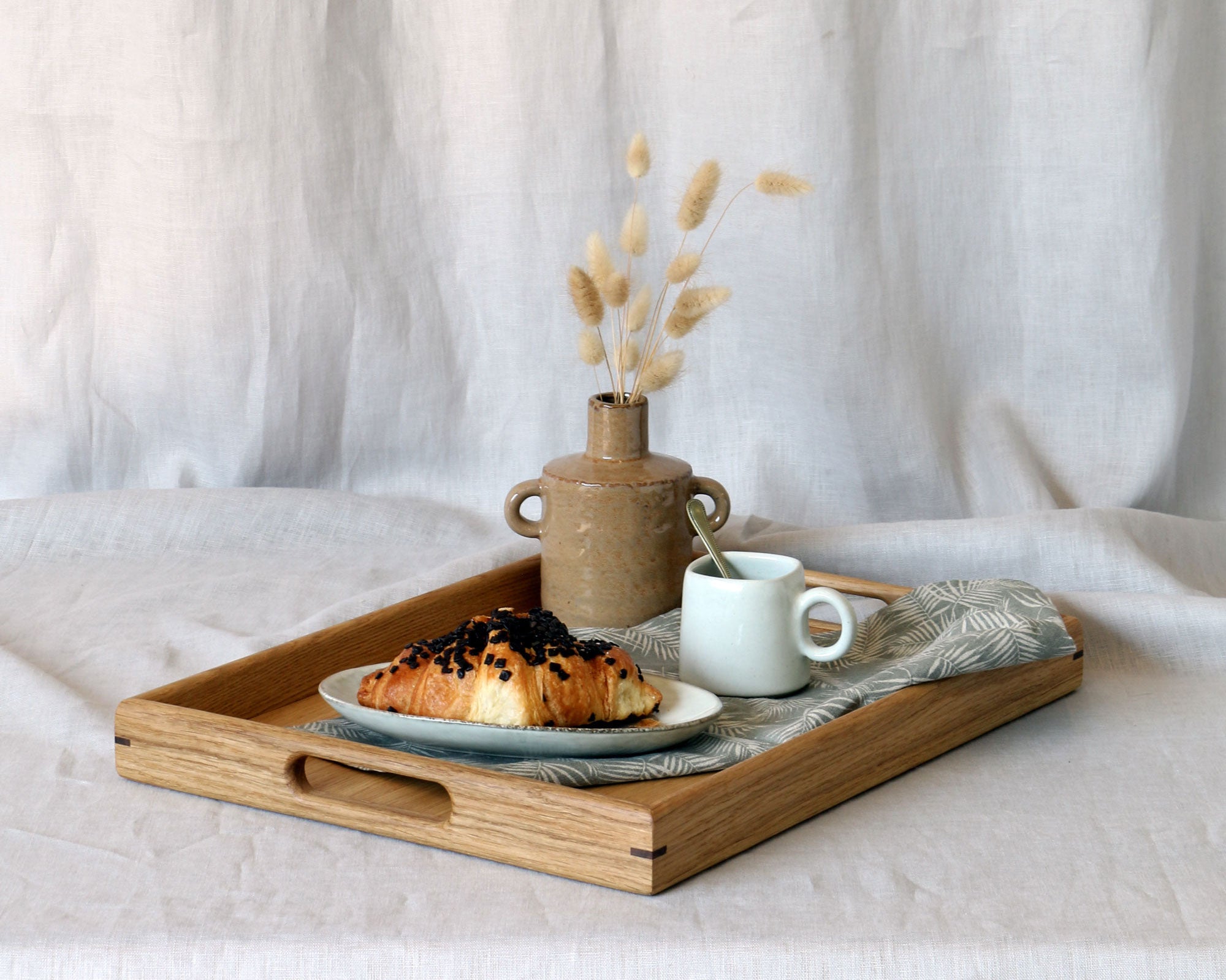 Breakfast-in-bed tray with handles, featuring a rustic ceramic vase, a cup of coffee, and a chocolate croissant on a linen napkin, set against a neutral fabric backdrop.