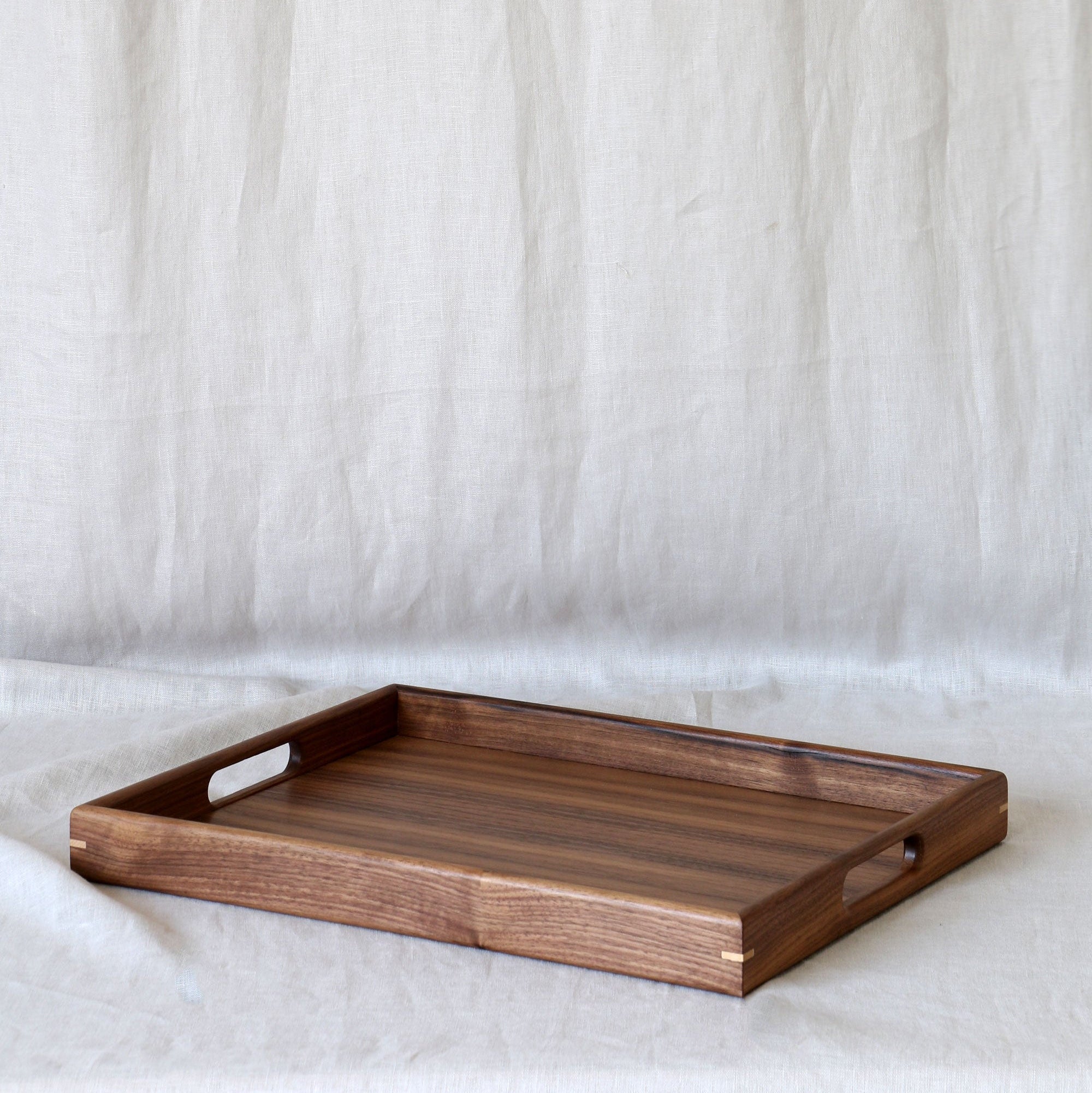 A rectangular wooden serving tray with cutout handles, shown on a draped off-white fabric backdrop.