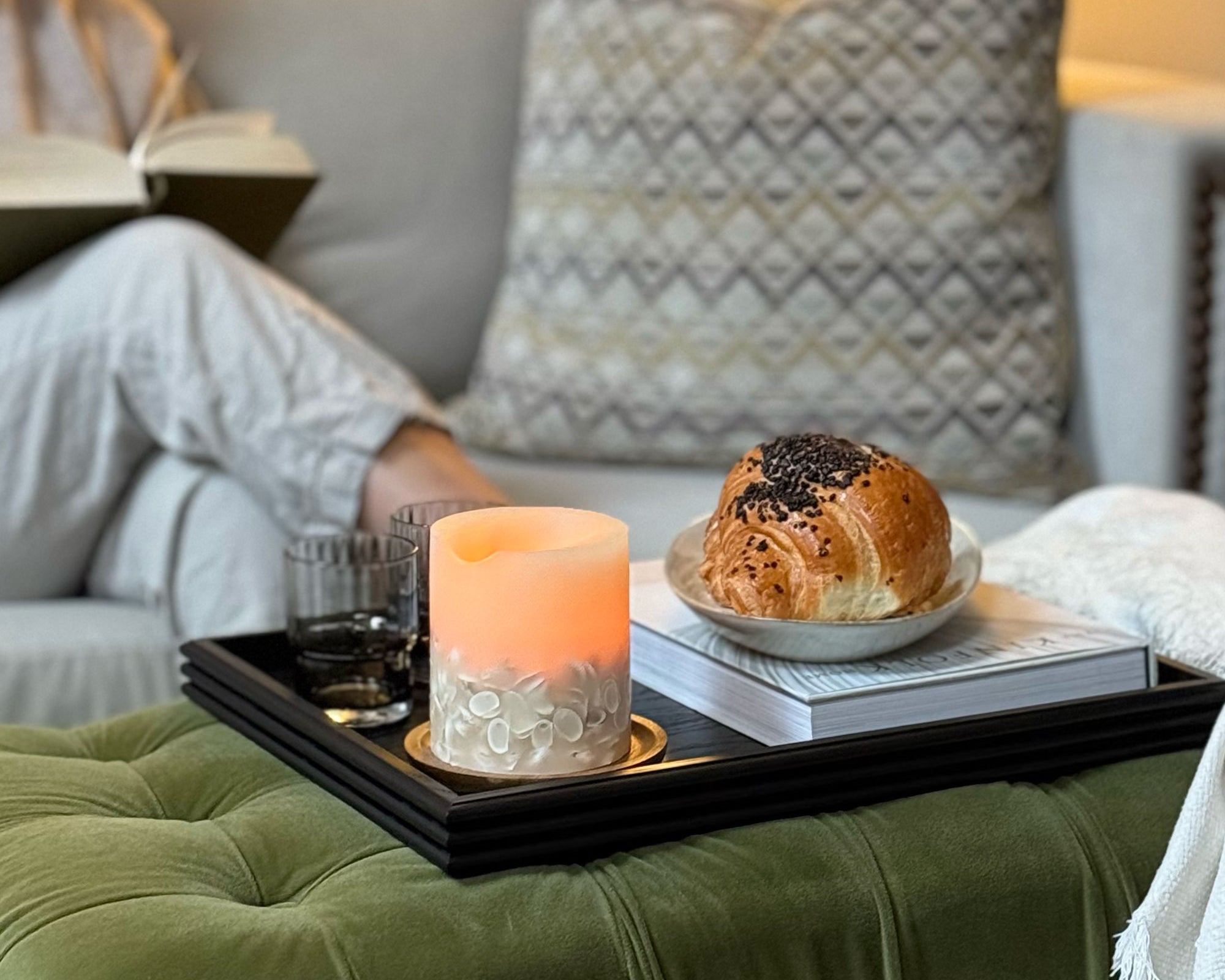 A wooden tray for a footstool, featuring a handcrafted black wood design with reeded edges, styled with a candle, glasses, and a croissant, creating a cozy and elegant living space.