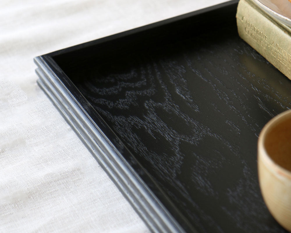 Close-up of a black wooden tray corner, showing the rich wood grain texture and reeded detailing along the edges, partially styled with a ceramic cup and book.