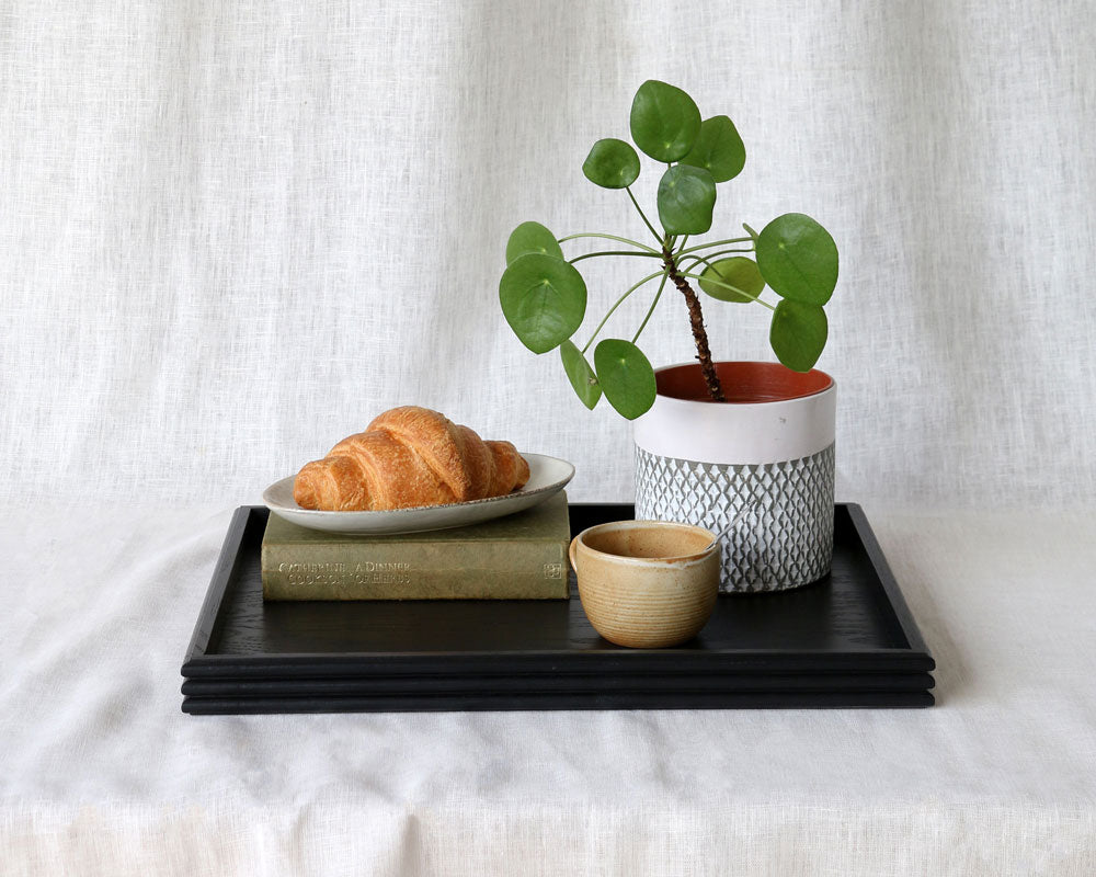 The black tray styled with a plate of croissants, a ceramic mug, a potted Pilea plant, and a hardcover book, placed on a white fabric surface.
