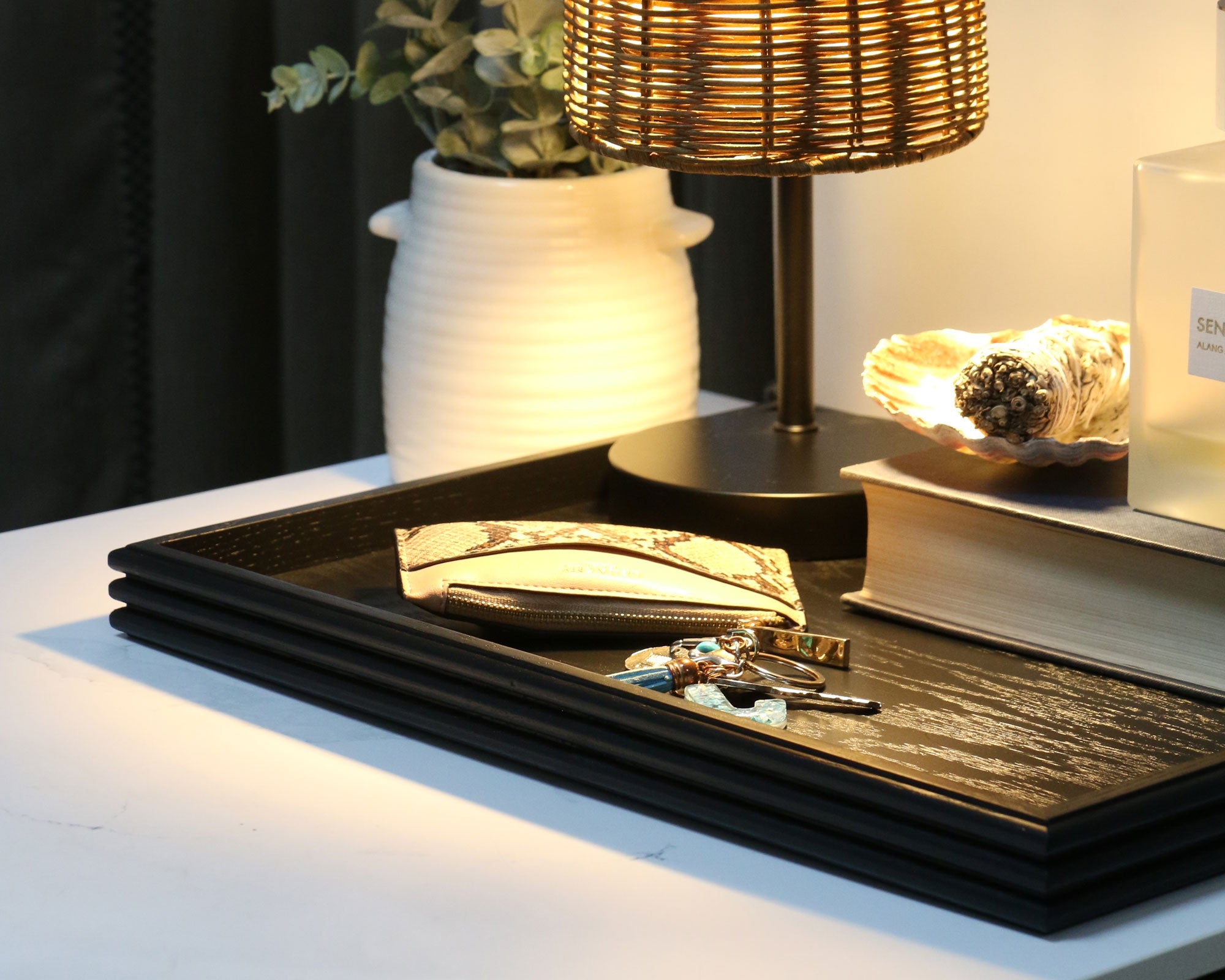 A decorative tray in black wood, styled on a modern nightstand with a woven lamp, a vase with greenery, and a sage bundle, adding warmth and sophistication to the space.