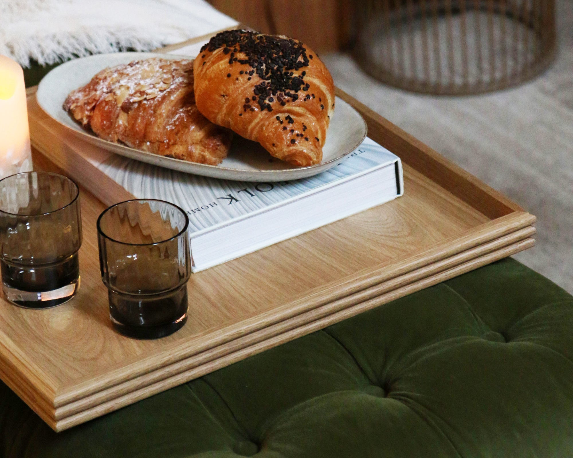 A coffee table tray in oak, neatly placed on a green tufted footstool, holding glasses, a croissant, and a book, adding warmth and functionality to the space.