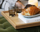 A decorative tray with a reeded wood design, styled with croissants, a book, and candles, offering a refined touch to a modern home aesthetic.