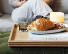 A wooden tray for a footstool, featuring a handcrafted oak design with reeded edges, elegantly styled with pastries, a book, and a candle in a cozy living room setting.