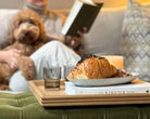 A handcrafted tray in natural oak, beautifully displayed with pastries, a candle, and glasses, set on a tufted ottoman, enhancing the ambiance of a cozy reading nook.