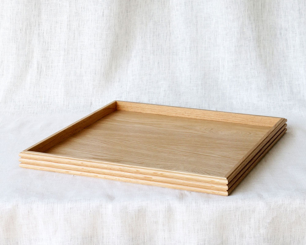 A square wooden tray made from oak, featuring subtle reeded detailing along the edges, placed on a soft white linen backdrop.