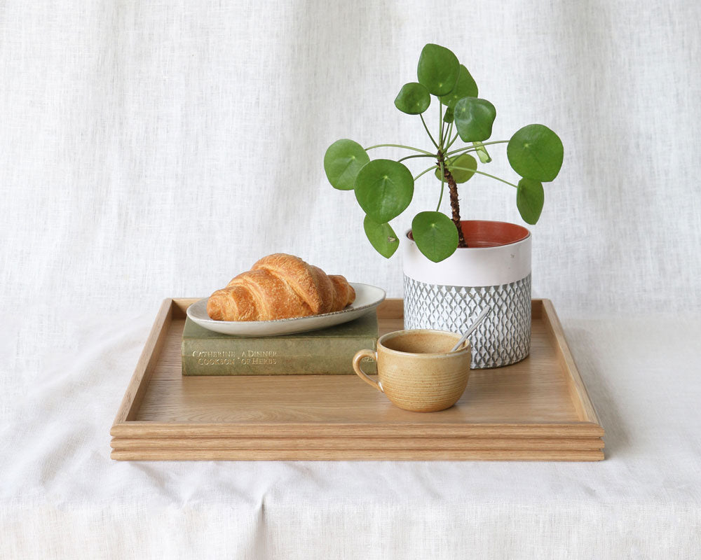 The oak tray styled with a croissant on a plate, a ceramic mug with a spoon, a potted Pilea plant, and a hardcover book, all resting on a white fabric surface.