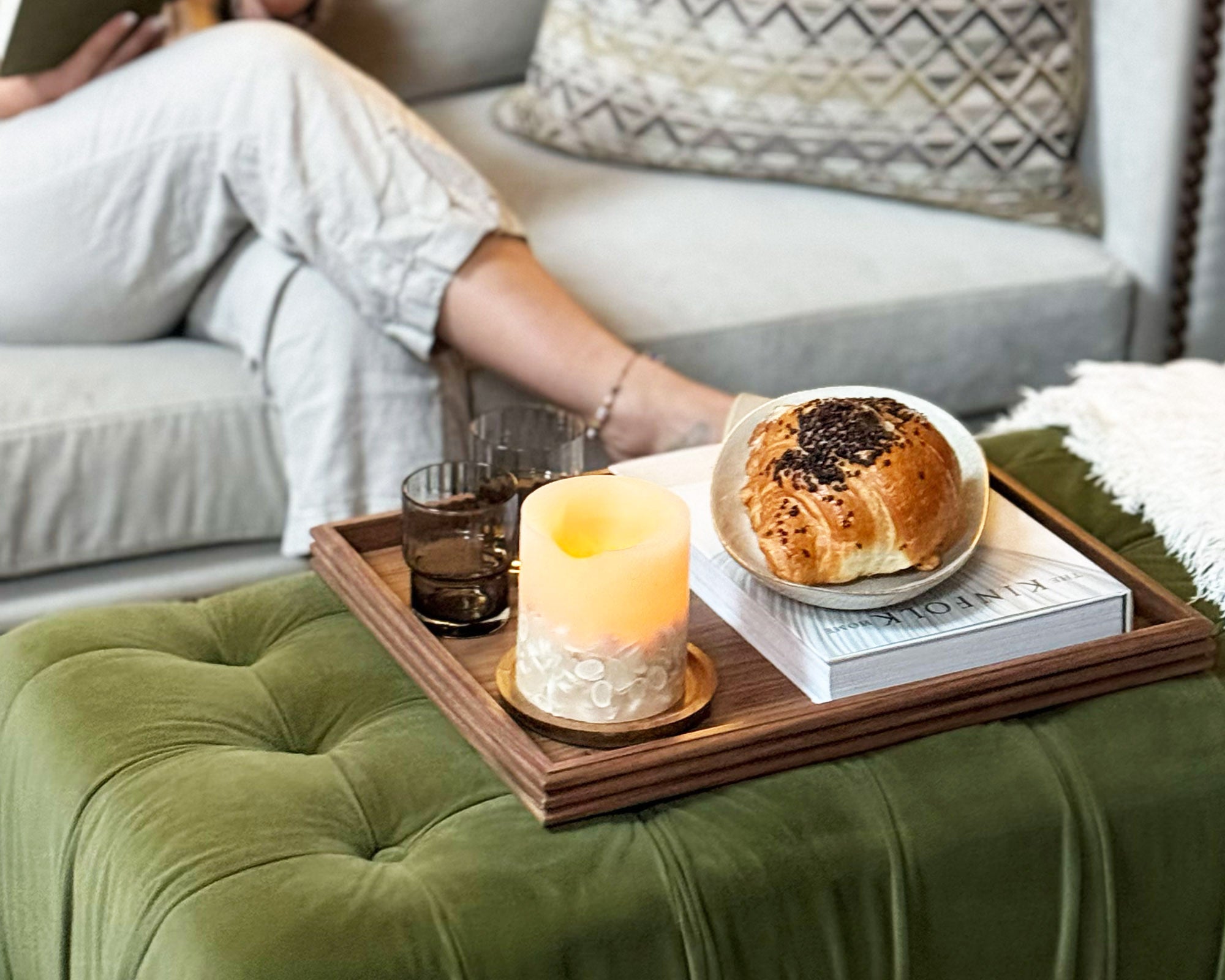 A decorative tray made of walnut, placed on a tufted ottoman, holding a croissant, book, and candle, creating a warm and inviting aesthetic.