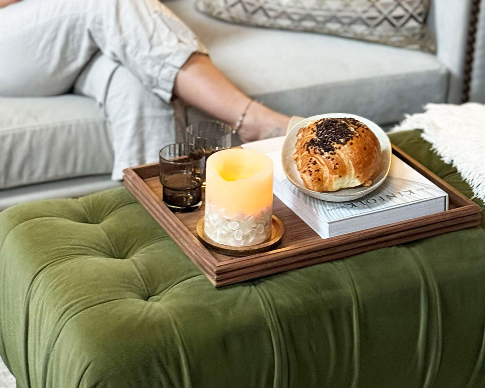 A decorative tray made of walnut, placed on a tufted ottoman, holding a croissant, book, and candle, creating a warm and inviting aesthetic.

