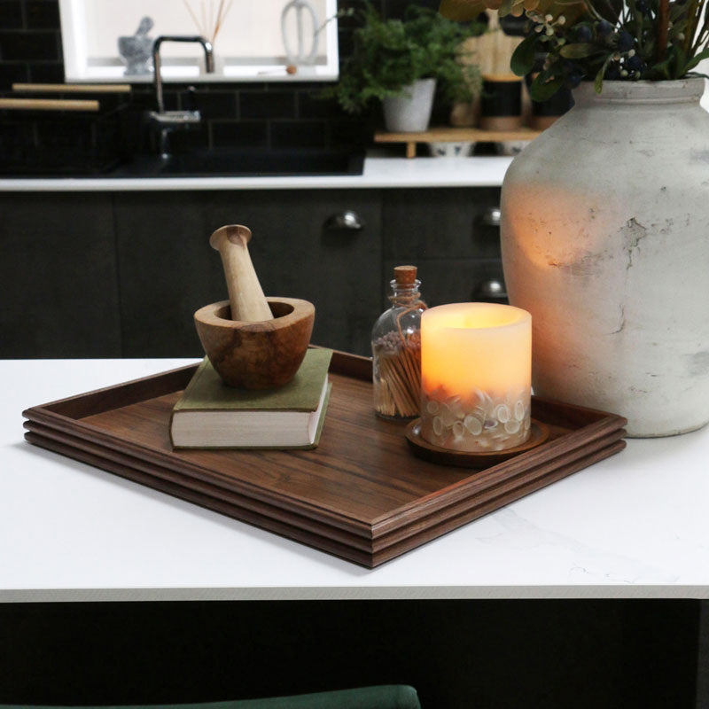 A handcrafted wooden tray styled on a kitchen counter, featuring a rich walnut finish, adorned with a candle, book, and decorative objects, blending functionality with aesthetic appeal.