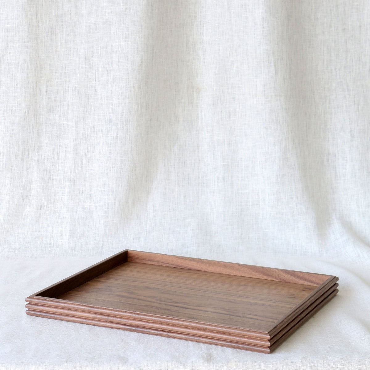 A rectangular wooden serving tray with a warm brown wood grain finish, displayed on a draped off-white linen fabric background.